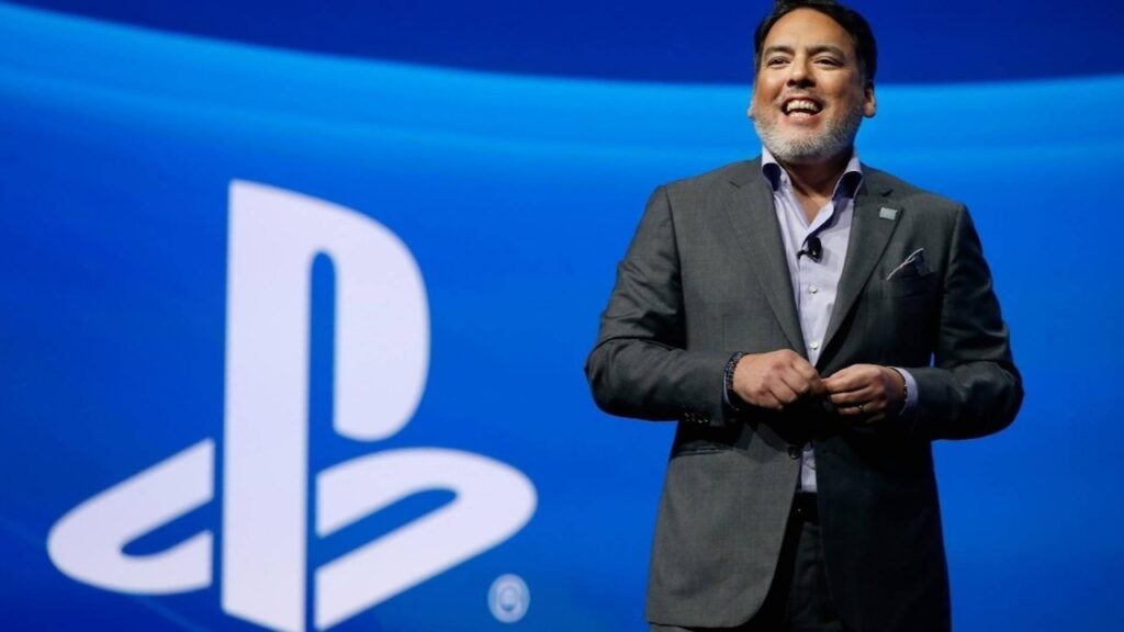 Shawn Layden sorridente con dietro il logo PlayStation