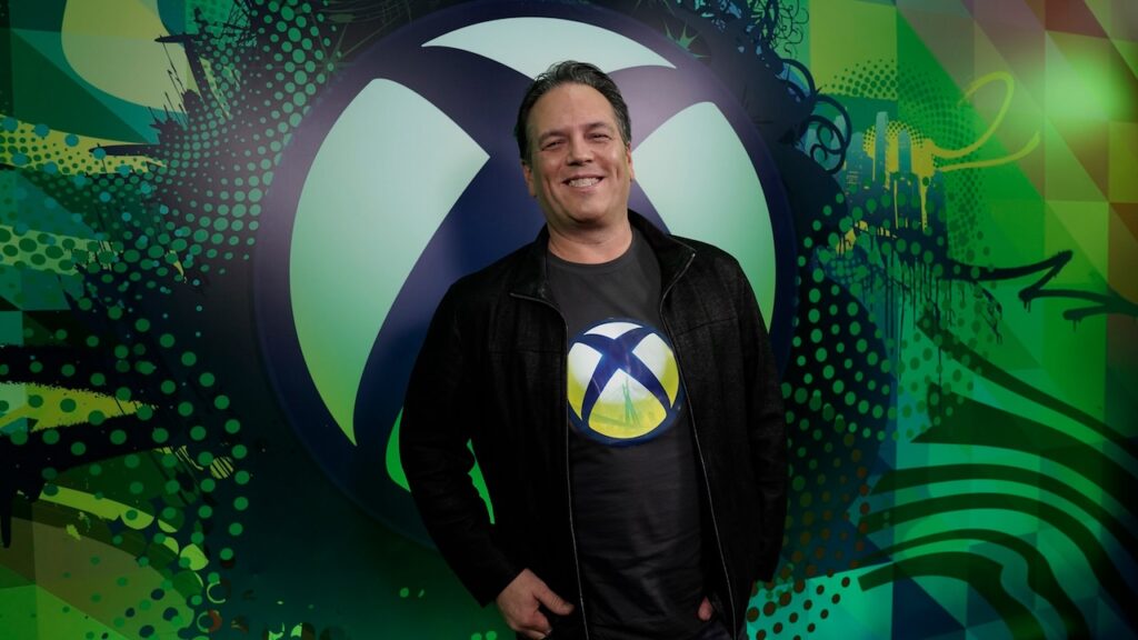 Phil Spencer sorridente con dietro il logo di Xbox