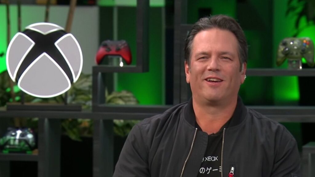 Phil Spencer sorridente con dietro il logo di Xbox